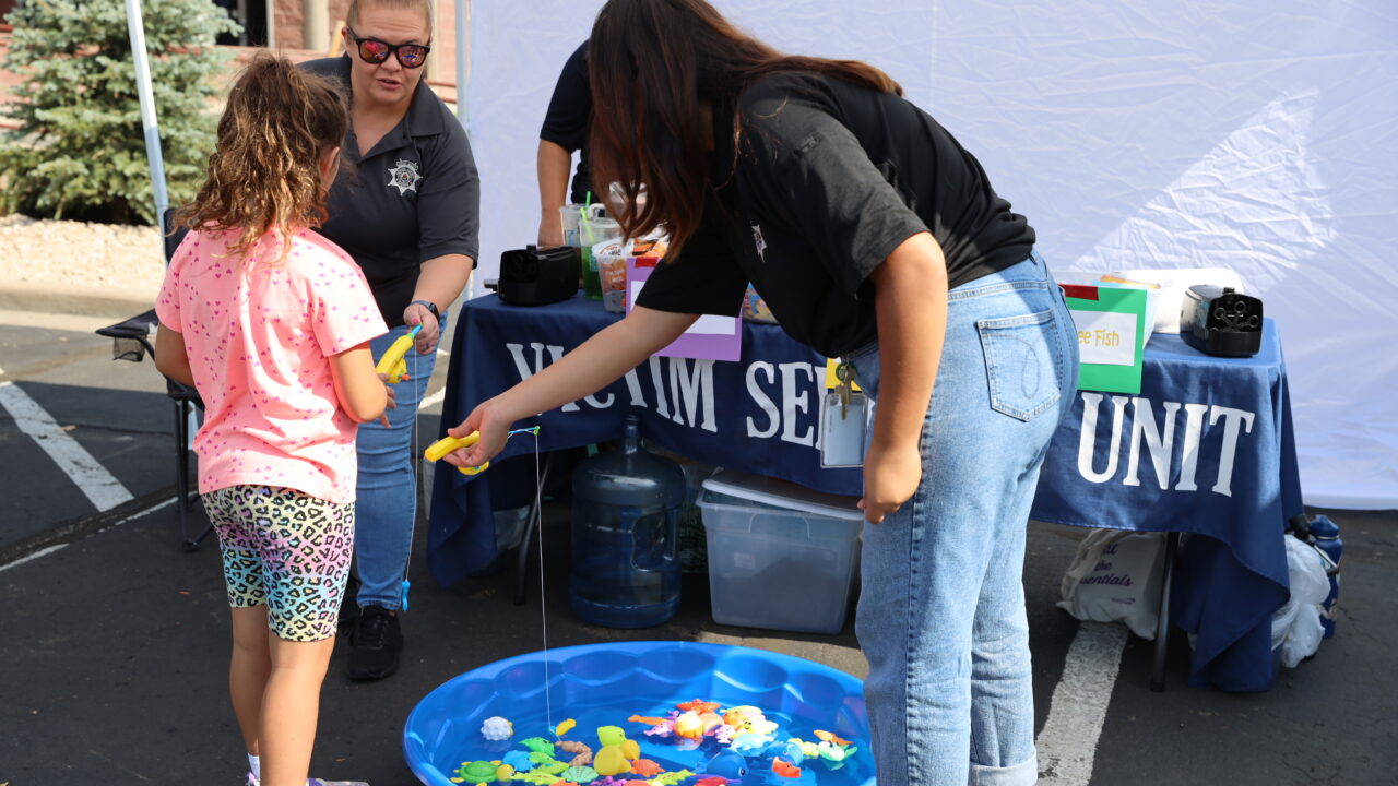 A woman assists a child using toy fishing rods beside a table labeled "Victim Services Unit." A small pool contains plastic fish.