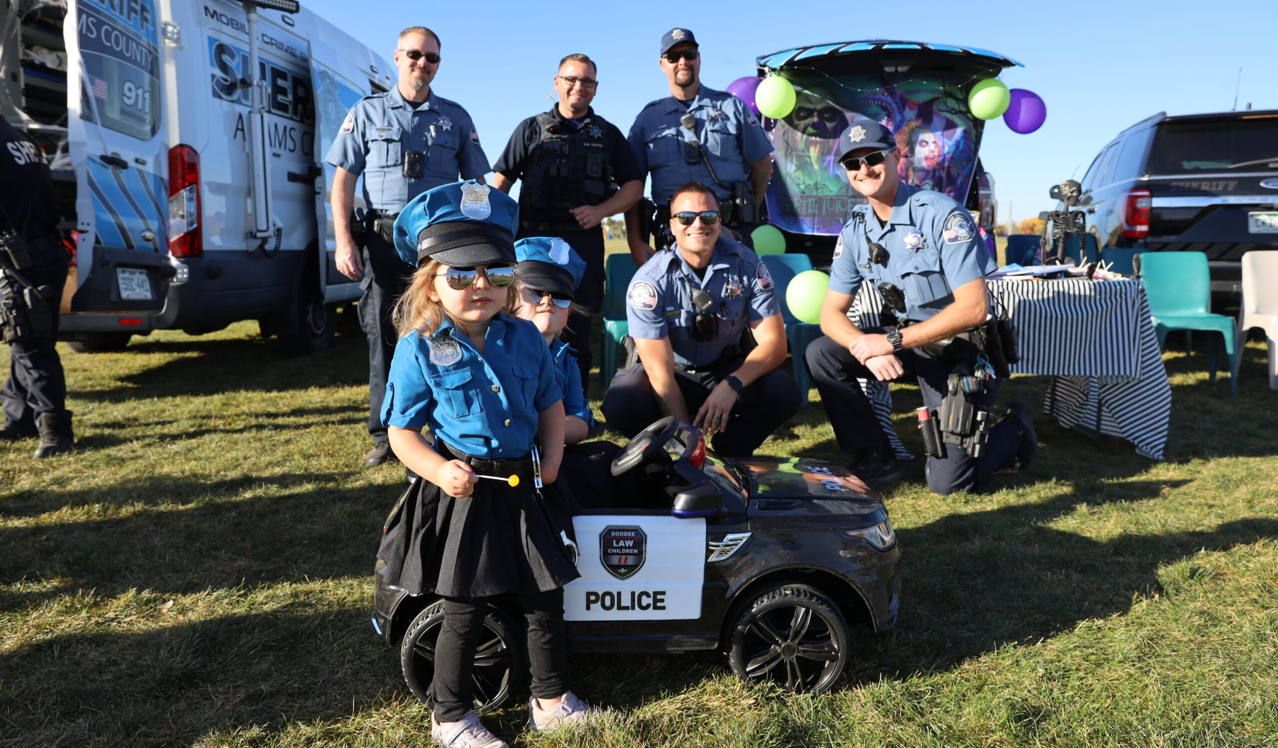 Children dressed as police officers pose with sheriff deputies, near sheriff vehicle at community event.