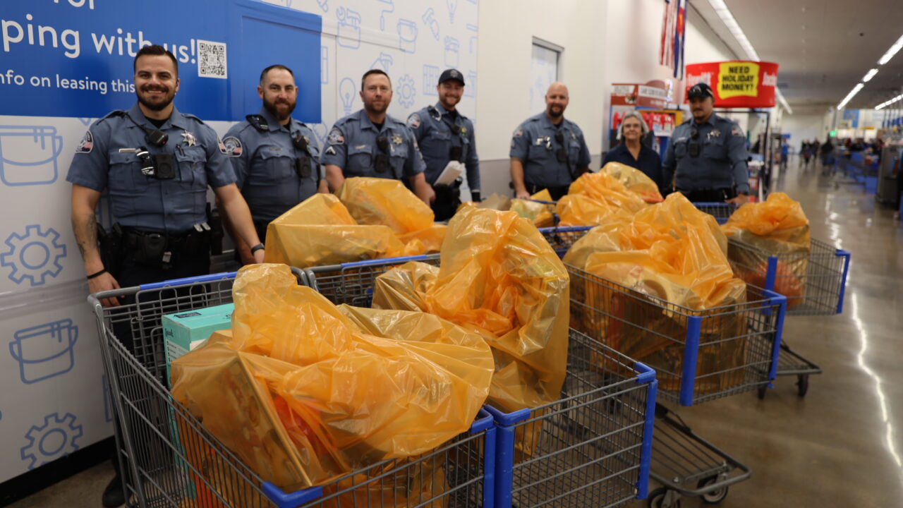 Officers in uniform standing behind grocery carts filled with yellow-bagged items, in a store with a wall sign reading "Thanks for shopping with us!"