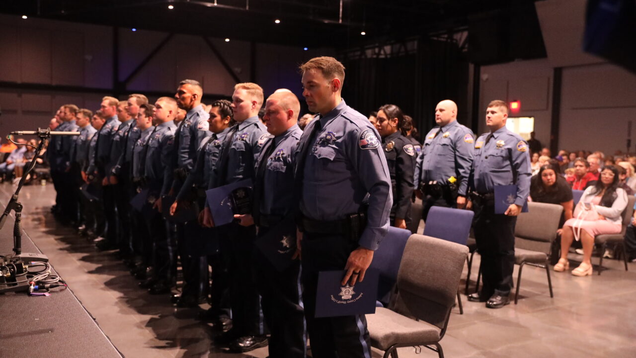 Graduates in uniform stand during a ceremony, holding certificates, with an audience in the background.