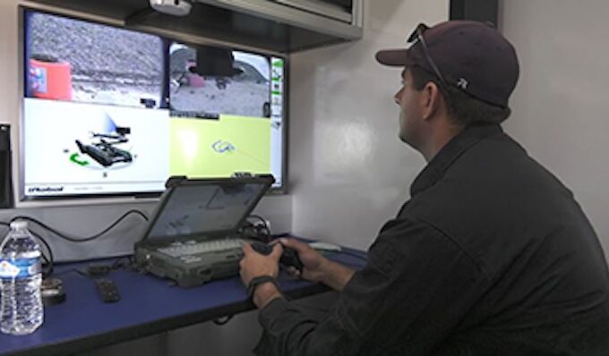 Adams County Sheriff monitors bomb squad drone footage on multiple screens, holding a remote control.