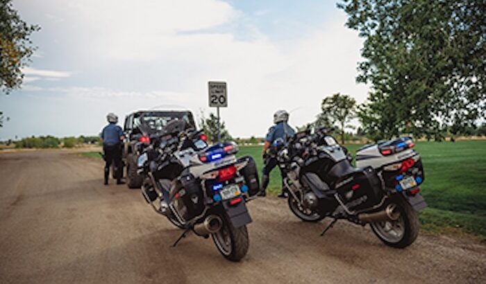 Adams County Sheriff traffic officers stand in front of their motorcycles near a parked vehicle on a rural road. Speed limit sign reads "Speed Limit 20".