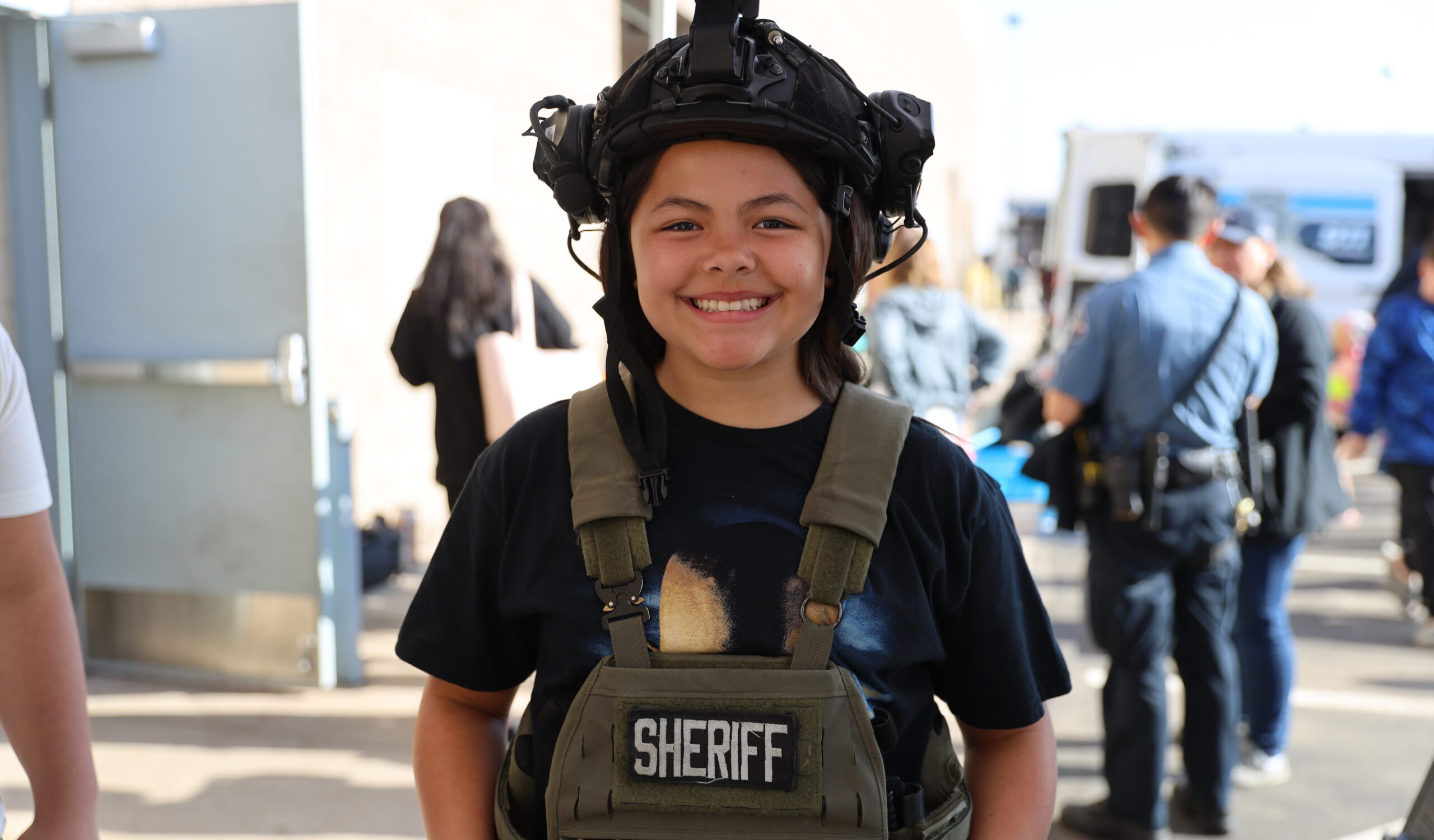 A young smiling person wearing a tactical helmet and ballistic vest at an outdoor event.