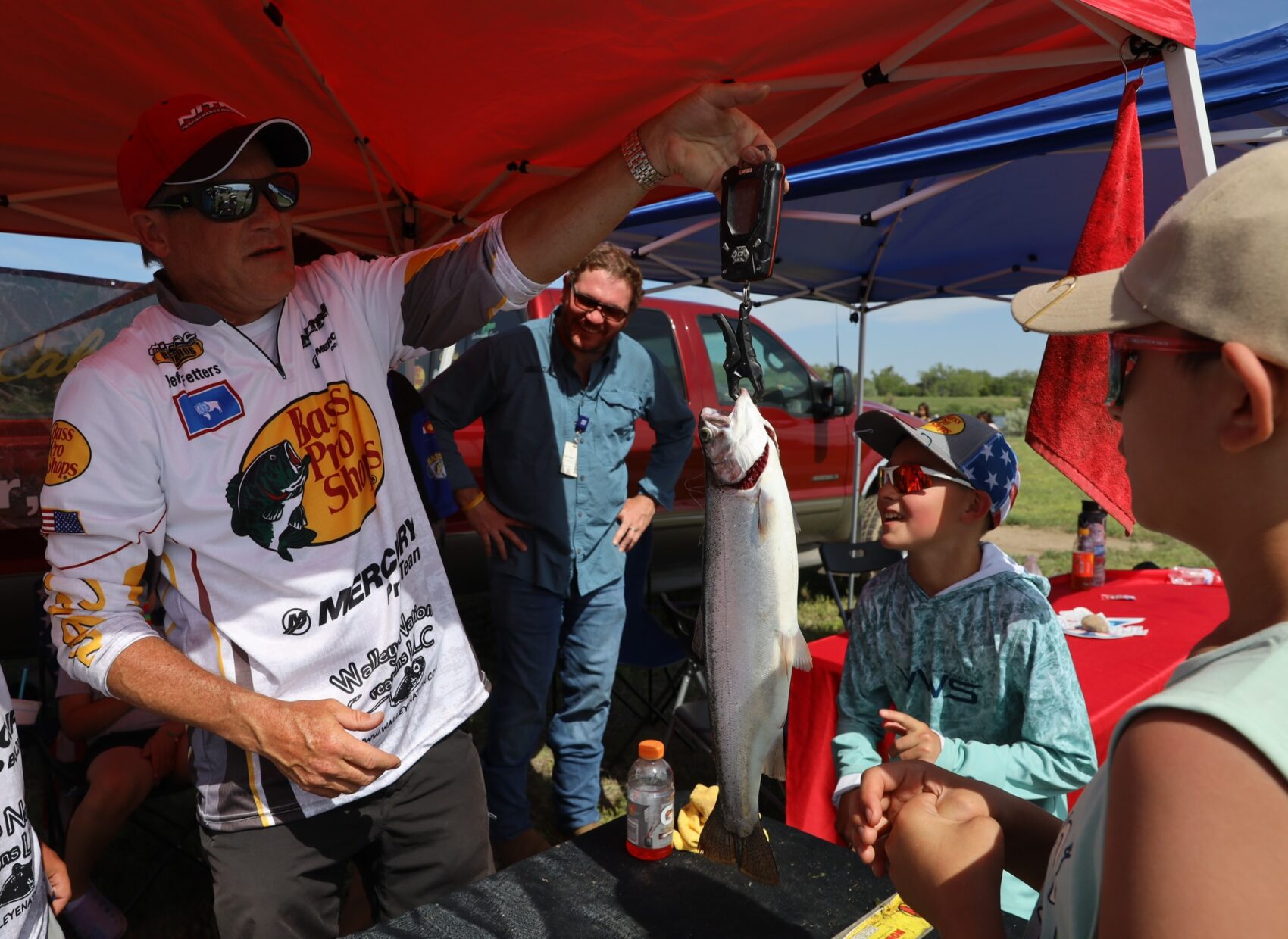 Man in a Bass Pro Shops jersey weighing a large fish on a scale as children watch during the Youth & Senior Fishing Festival.