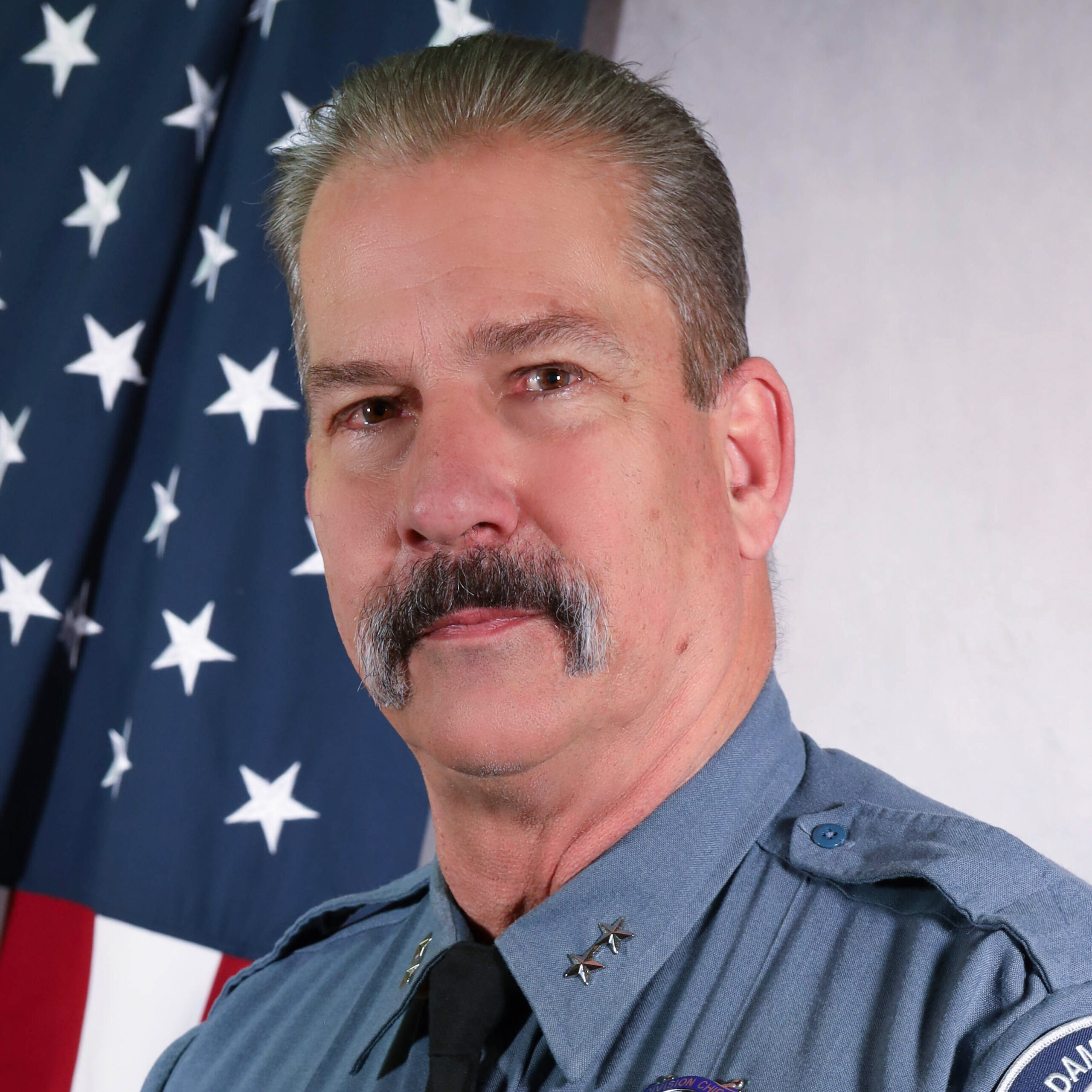 Portrait of Division Chief John Bungartz in uniform, smiling in front of an American flag. He wears a badge and tie, with the Adams County Sheriff’s Office patch on his left sleeve.
