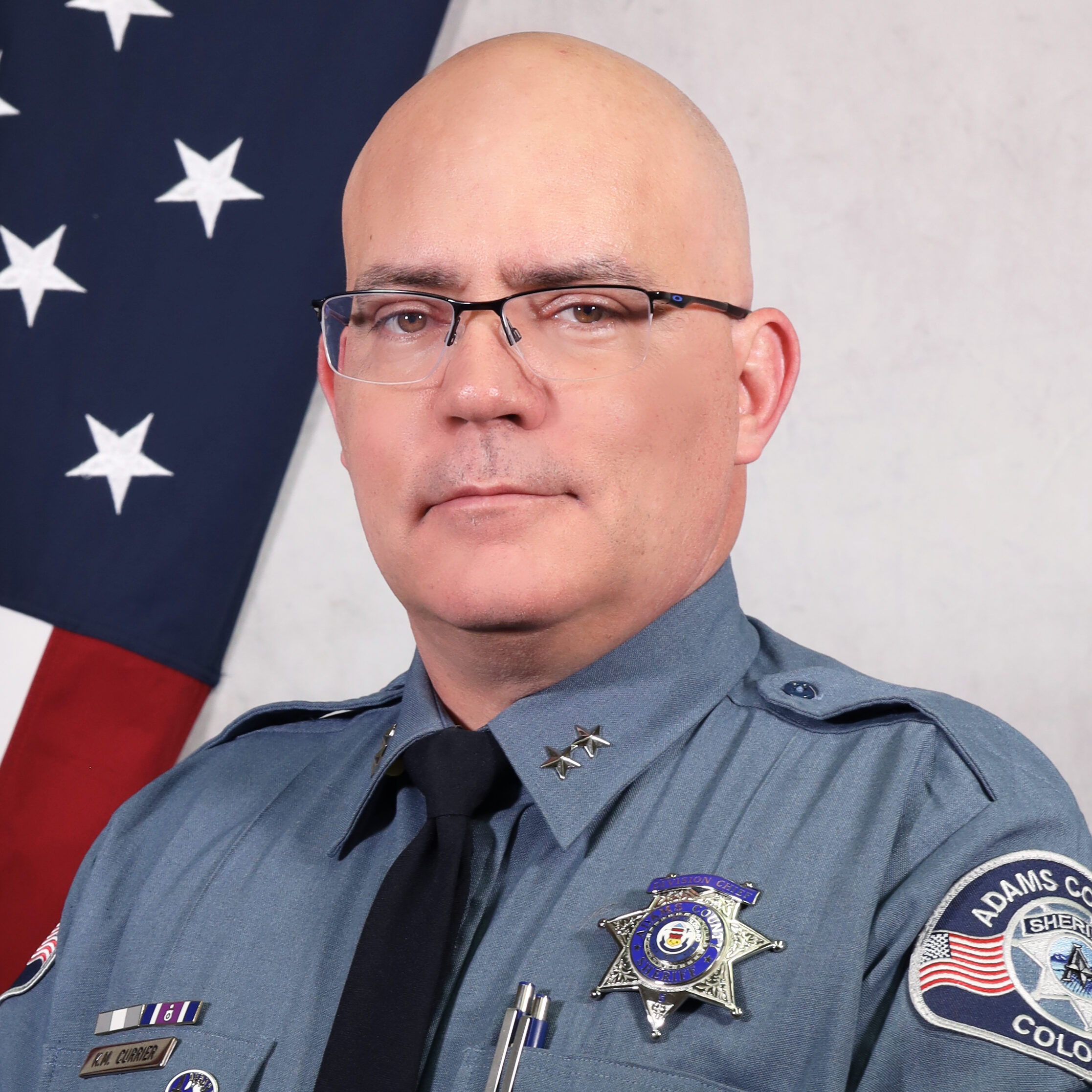 Portrait of Interim Undersheriff Kevin Currier in uniform, smiling in front of an American flag. He wears a badge, service pins, and a black tie, with the Adams County Sheriff’s Office patch visible on his left sleeve.