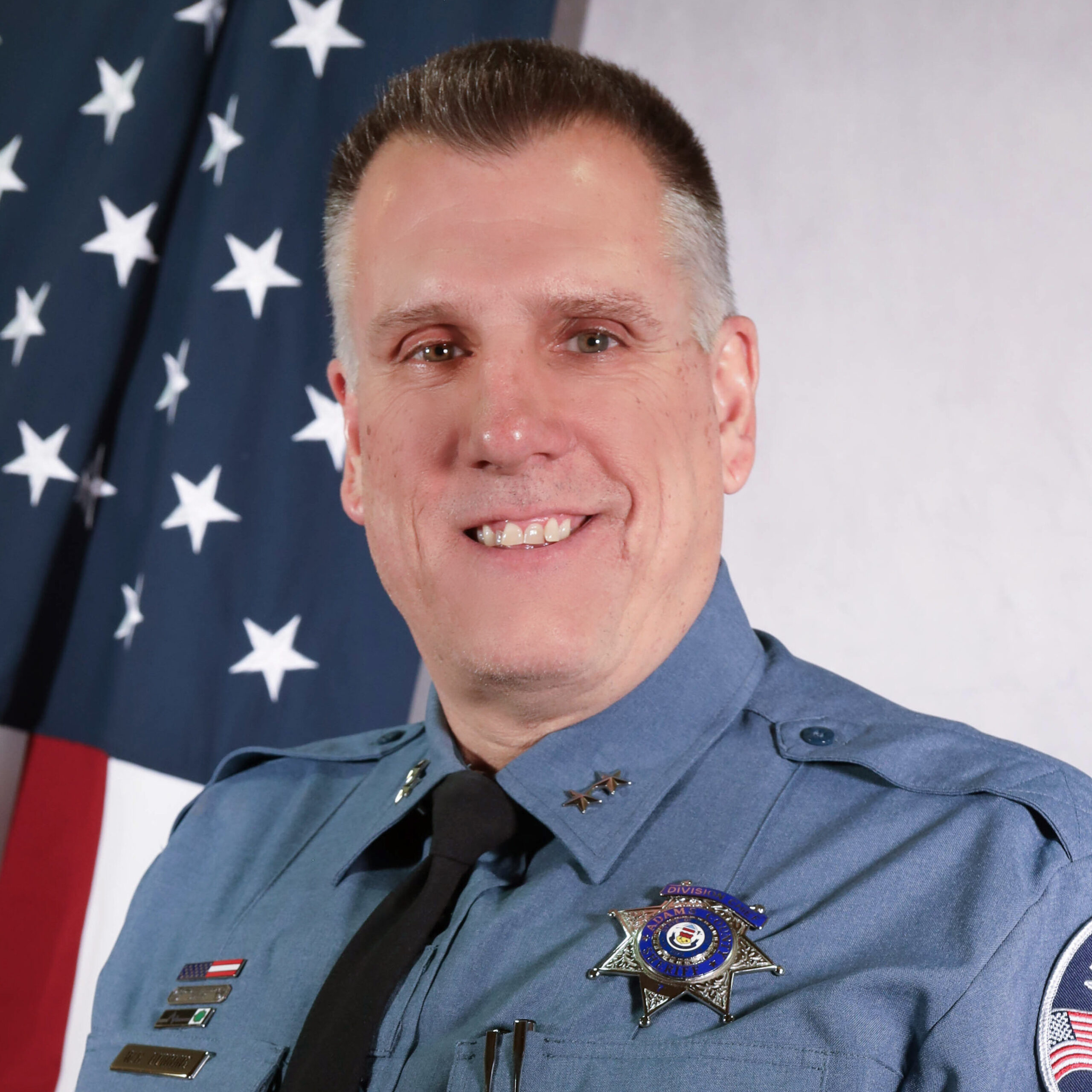 Portrait of Jail Division Chief Bill Dunning in uniform, smiling in front of an American flag. He wears a badge, service pins, and a black tie, with the Adams County Sheriff’s Office patch on his left sleeve.