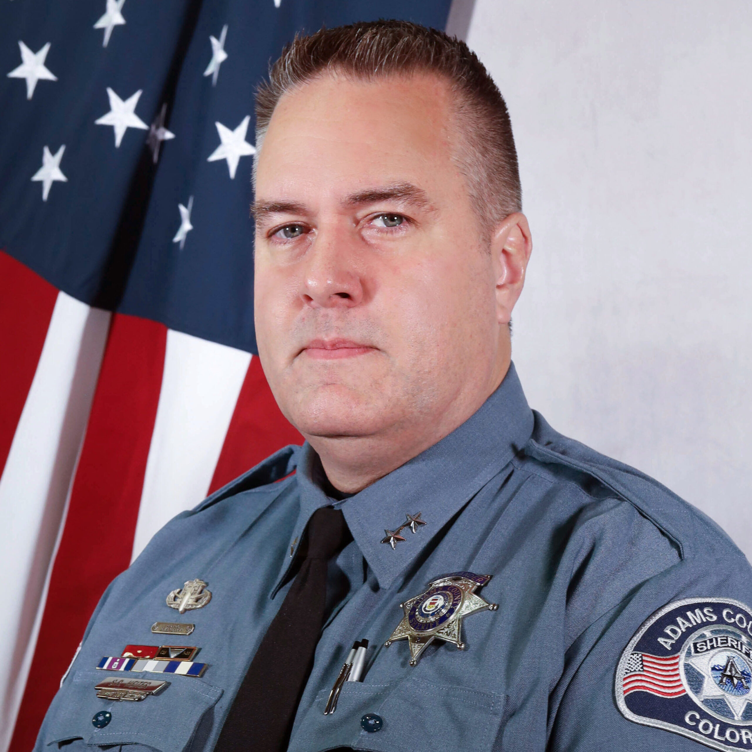 Portrait of Training Division Chief Shane Heiter in uniform, standing in front of an American flag. He wears a badge, various service pins, and a black tie. The Adams County Sheriff’s Office patch is visible on his left sleeve.