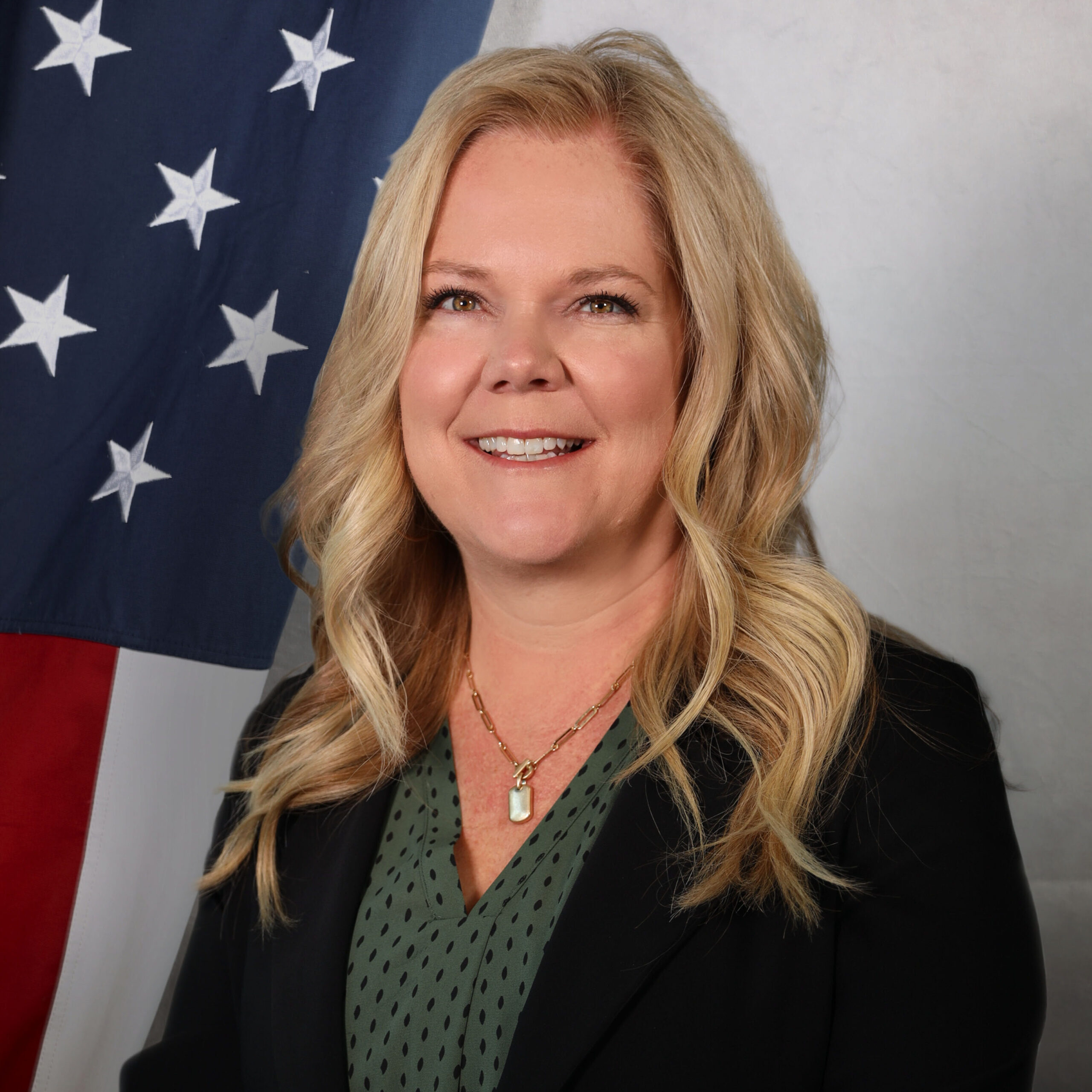 Portrait of Jennifer Baessler, HR Director at the Adams County Sheriff’s Office, smiling in front of an American flag, wearing a black blazer and green blouse.