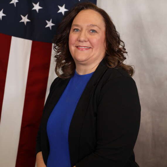 Recruiter Lacrue, Adams County Sheriff's Office recruiter, smiling, standing in front of an American flag backdrop.