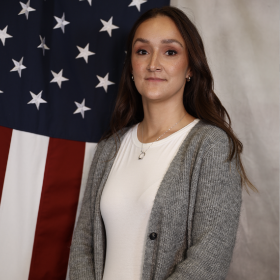 Thya Salazar, Adams County Sheriff's Office recruiter, standing in front of an American flag, smiling slightly.