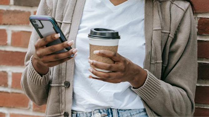 Person leaning against a brick wall, holding a smartphone and a coffee cup.