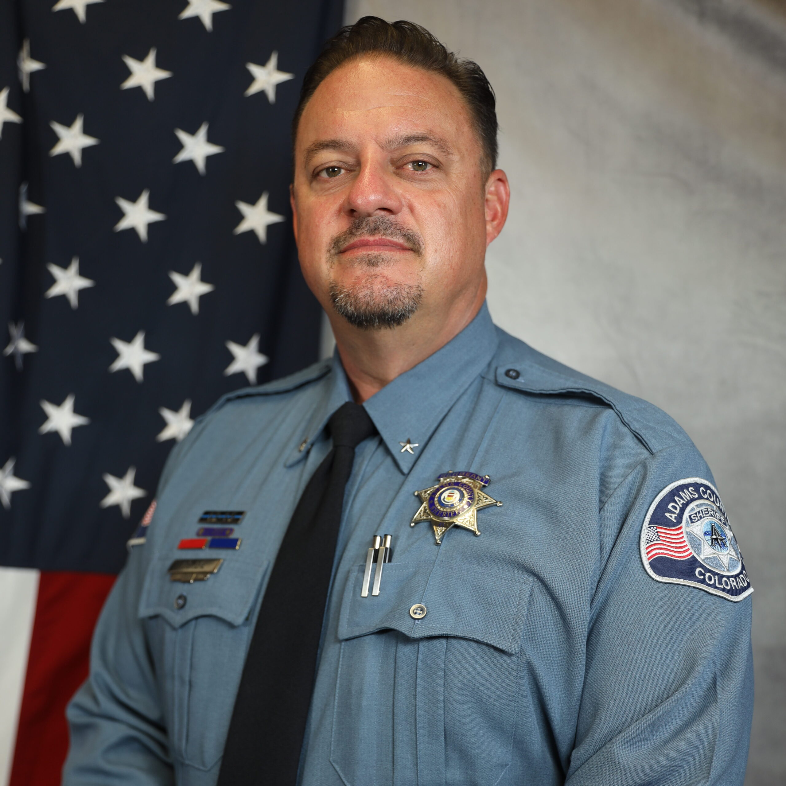 Man in Adams County sheriff uniform stands in front of American flag