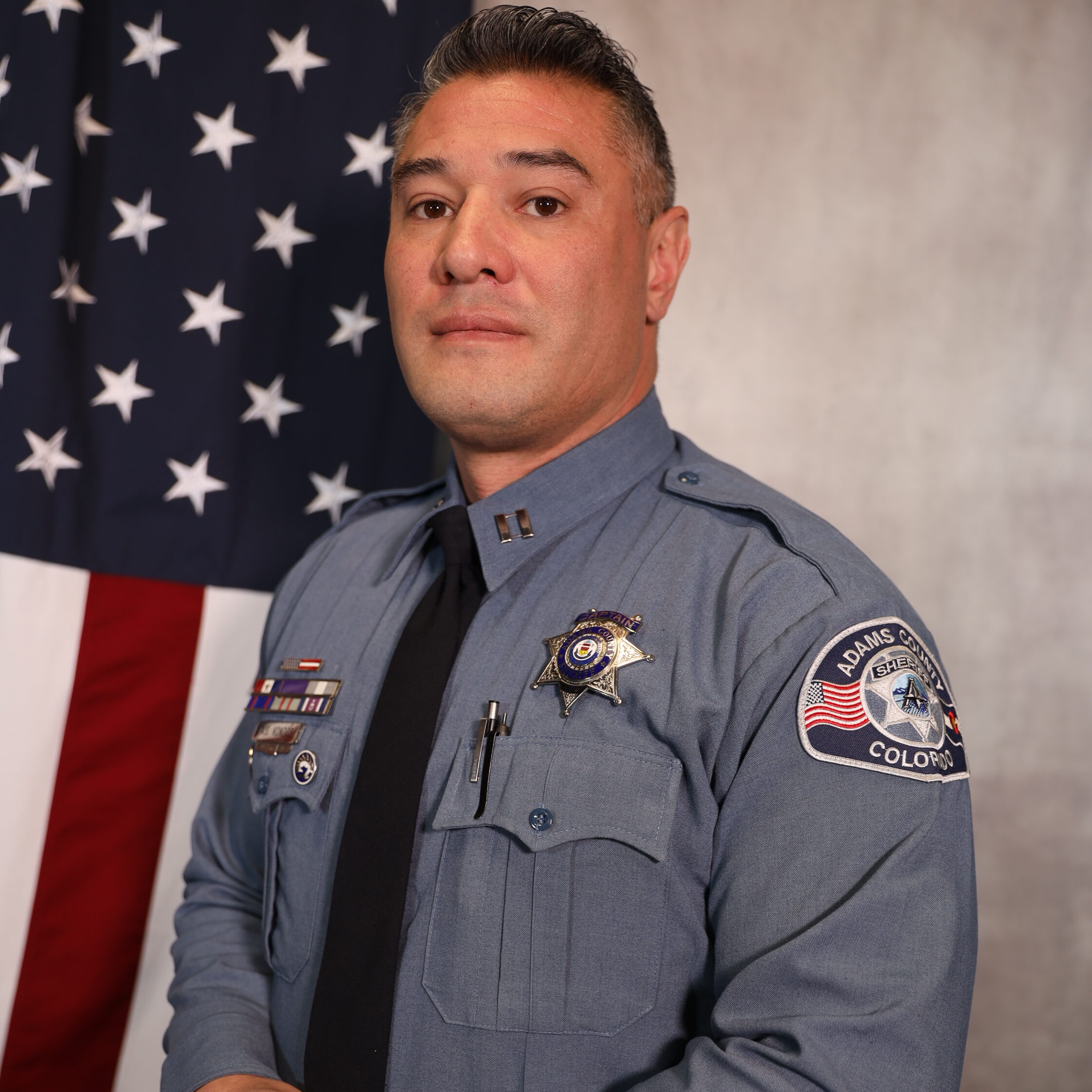 Kondos-Alex, in uniform, stands confidently beside the American flag, displaying law enforcement insignia.
