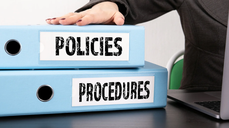 Hand on binders labeled "Policies" and "Procedures" next to a laptop